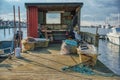 A rusty anchor with a light blue rope tied up on a wooden pier. In the background a fisherman hut with lots of fishing tools