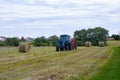 Rybushkino village, Russia - July 1, 2020: Blue tractor collects large round rolls of hay from dry clover grass. Haymaking.