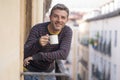 30s or 40s attractive and happy man at home balcony relaxed and cheerful enjoying cup of coffee looking to city street smiling and