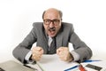 60s bald senior office boss man furious and angry gesturing upset and mad sitting on desk with paperwork in business and job prob Royalty Free Stock Photo