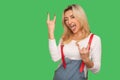 That`s crazy! Portrait of delighted adult woman in stylish denim overalls showing rock and roll gesture