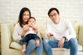 30s happy Asian family are watching TV and smiling while sitting on couch at home. Father is holding remote control in Royalty Free Stock Photo