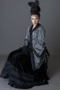 A Victorian woman wearing a striped silk polonaise and a black skirt and sitting down against a studio backdrop Royalty Free Stock Photo