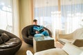 Young man sitting in a chair reading a book in a comfortable library room near huge windows. Royalty Free Stock Photo