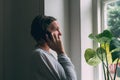 Sad alone woman talking on mobile phone while looking out the window of living room. Casual mid-adult brunette with serious facial Royalty Free Stock Photo