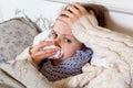 Sad alone young woman in white sweater and blue scarf feeling cold sick and resting home in bed. cleaning her nose, holding her Royalty Free Stock Photo