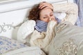 Sad alone young woman in white sweater and blue scarf feeling cold sick and resting home in bed. cleaning her nose, holding her