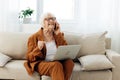 a sad, angry elderly woman in a brown suit is sitting with a laptop on her lap on a cozy sofa working from home and Royalty Free Stock Photo