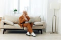 a sad, angry elderly woman in a brown suit is sitting with a laptop on her lap on a cozy sofa working from home and Royalty Free Stock Photo