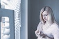 Sad apathetic lonely woman standing near the window and texting messages in smartphone at home or hotel, divorce, depression and Royalty Free Stock Photo