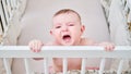 A sad baby is crying in a crib, holding onto the handrails. Funny child in bed, age six months