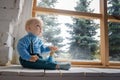 Sad, but beautiful blue-eyed baby sitting on the windowsill and not looking at the camera Royalty Free Stock Photo