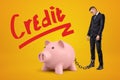 Sad businessman chained to pink piggy bank with `Credit` sign above on yellow background.