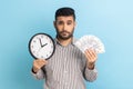 Sad businessman holding big fan of dollar banknotes and wall clock, time is money, looking at camera Royalty Free Stock Photo