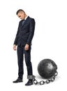 A sad businessman looks down while shackled to a large iron ball with a chain to his ankle.