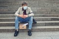 Sad businessman in a medical mask sitting on the stairs outdoors with a box of documents. A desperate male office worker