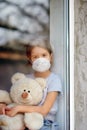 Sad Child, little girl in mask with teddy bear looking from window, coronavirus quarantine Royalty Free Stock Photo