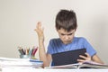 Sad confused child with tablet computer sitting at table with books notebooks