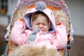 Sad crying little beautiful baby girl sitting in the pram or stroller on autumn day. Unhappy tired and exhausted child