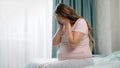 Sad crying pregnant woman suffering from depression sitting on bed and holding her head. Concept of maternal and