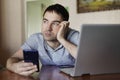 Sad cute young man holding a smartphone in his hand a man working on a laptop remotely. Fatigue from quarantine, self-isolation Royalty Free Stock Photo