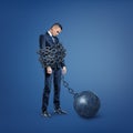 A sad disappointed businessman stands bound by a wide metal chain which is locked to an iron ball.