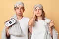 Sad displeased couple man and woman in sleep eye mask wrapped in blanket holding coffee cup and alarm isolated over beige Royalty Free Stock Photo