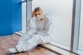 Sad doctor holding hand over head and sitting on the floor near window in hospital because of coronavirus epidemy. Portrait of ups