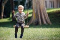A sad, dreaming little boy swings in a city park under a big tree Royalty Free Stock Photo