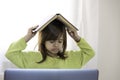 Sad elementary schooler girl studying at home with a book on her head because she is tired and angry - Little girl studying online