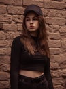 Sad emotional young woman looking unhappy near the red brick wall on the street background in black style clothing and cool cap. Royalty Free Stock Photo