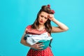 Sad exhausted pinup woman in retro outfit holding clothes for washing or ironing on blue studio background Royalty Free Stock Photo