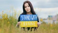 Sad female ukrainian girl migrant woman upset lady posing in field outdoor colorful written cardboard stand with Ukraine