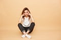 Sad girl dressed in white t-shirt, jeans and white socks sits on the floor on the beige background in the studio