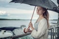 Sad girl hiding under a black umbrella from the rain, standing on the banks of the river. Curls of hair develop in the wind. Storm