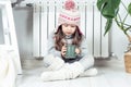 Sad girl in woolen hat, socks and pullover hold hot drink and sit with crossed legs near heating convector, calorifer