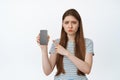 Sad and gloomy girl pointing finger at mobile phone screen, looking disappointed and upset. Young woman complains at Royalty Free Stock Photo