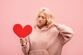 Sad and heart broken young lady holding red paper heart and showing thumb down, posing over pink studio background Royalty Free Stock Photo