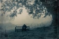 A sad hooded man sitting on a bench in the countryside looking down from the camera on a misty, moody day. With a grunge, vintage,