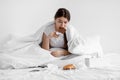 Sad hungry european millennial female eating big chocolate bar sitting on white bed with fast food in bedroom