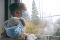 Sad illness child on home quarantine. Boy and his teddy bear both in protective medical masks sits on windowsill and looks out
