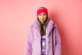 Sad and indecisive asian senior woman in fashionable faux fur coat looking left and sulking upset, standing against pink