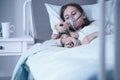 Sad kid with cystic fibrosis lying in a hospital bed with oxygen mask and plush toy