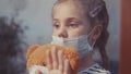 sad kid girl in protective medical mask by the window looks out. stay home coronavirus teddy bear concept. sad kid in Royalty Free Stock Photo