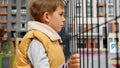 Sad little boy holding metal fence on public playground and looking through. Child depression, problems with bullying, victim in