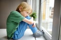 Sad little boy is sitting near window and watching street. Lonely at home. No friends, no siblings. One baby in family. Bad Royalty Free Stock Photo