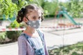 Portrait of a little girl in a protective mask on the background of a playground, fenced with a prohibition tape