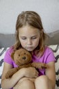 a sad little girl sits on the bed at home, hugs a teddy bear and worries. The concept of resentment and misunderstanding