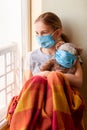 Sad little girl with teddy bear toy both in protective masks sitting on the window at home . Coronavirus quarantine Royalty Free Stock Photo