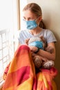 Sad little girl with teddy bear toy both in protective masks sitting on the window at home . Coronavirus quarantine Royalty Free Stock Photo